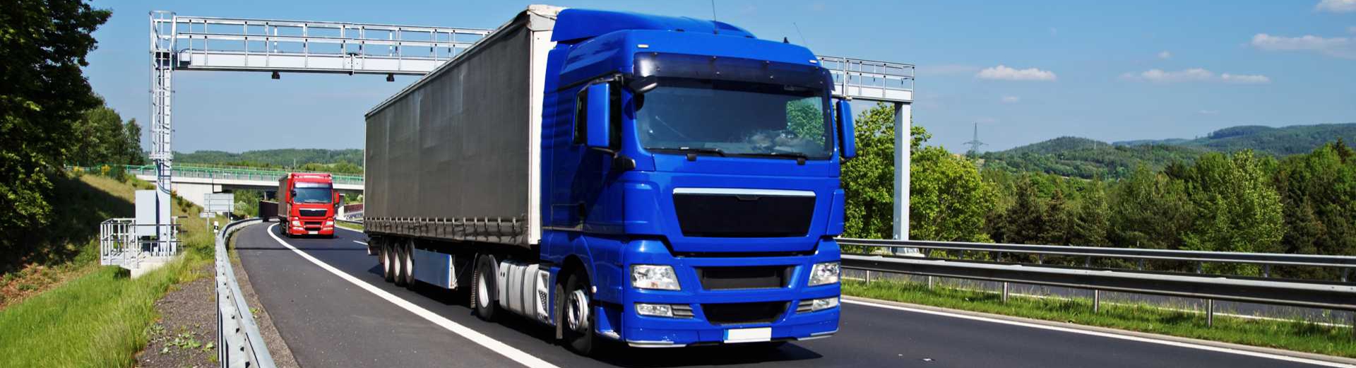 Blue truck passing through the electronic toll gates on the highway in a wooded landscape. Red truck, bridge and forested mountains in the background. White clouds in the blue sky.